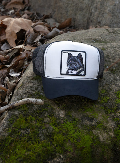 DogKingdom black and white trucker hat with Akita embroidery on a mossy rock surrounded by leaves