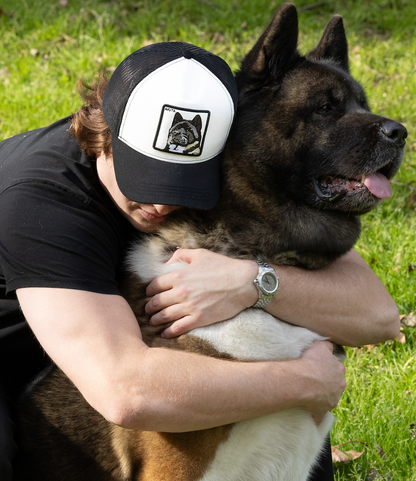 Person hugging a big Akita dog while wearing a DogKingdom trucker hat outdoors